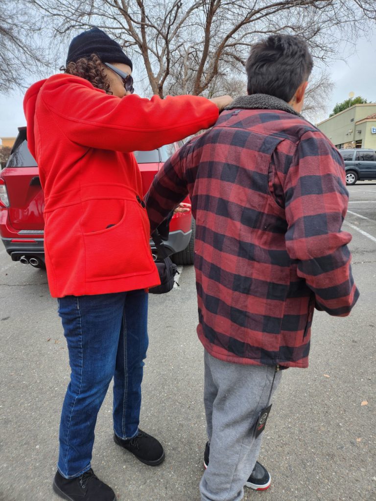 A Hart to Hart 4 U team member helps a young man put on a brand-new, warm flannel jacket in a parking lot.